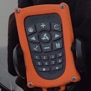 May include: Orange remote control with a black keypad featuring a variety of buttons, including directional arrows, a plus sign, a minus sign, and numbers 1 through 8. The remote control has a black strap attached to it.