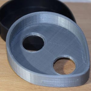 May include: A gray, 3D-printed object with two circular holes and a rounded shape, possibly a component or part. A black, circular object is partially visible in the background. The objects are on a wooden surface.