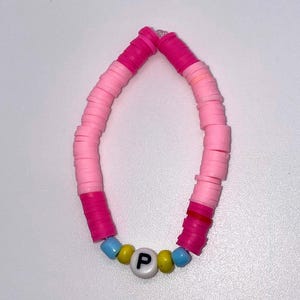 May include: A handmade bracelet featuring pink, light pink, and fuchsia flat disc beads. The bracelet includes a section of blue and yellow beads with a white bead displaying the letter "P". This colorful accessory is perfect for everyday wear.