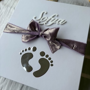 May include: A white gift box with a silver bow and baby footprints. The name "Sofia" is written in cursive. A purple ribbon with gold accents is tied around the box. A wooden hairbrush is visible in the background.