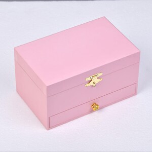 May include: A pink wooden jewelry box with a gold latch and a drawer. The box has a smooth, polished finish.