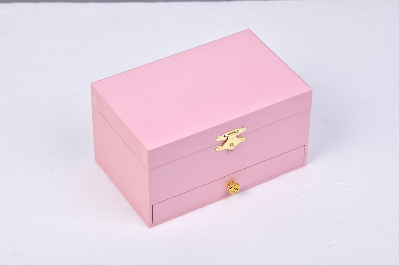 May include: A pink wooden jewelry box with a gold latch and a drawer. The box has a smooth, polished finish.