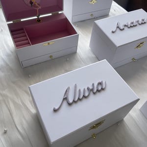 May include: White wooden music boxes with personalized names in cursive script. The boxes have a drawer and a ballerina figurine inside.