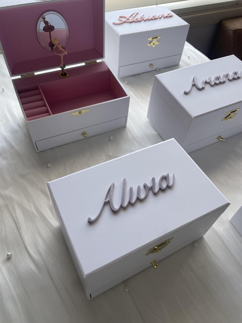 May include: White wooden music boxes with personalized names in cursive script. The boxes have a drawer and a ballerina figurine inside.