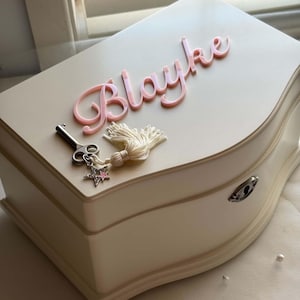 May include: A cream-coloured jewellery box with a curved front and a silver keyhole. The top features the name Blayke in raised pink script lettering, accompanied by a silver key charm with a white tassel and a small star-shaped pendant.