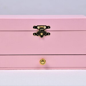 May include: A pink wooden jewelry box with a gold latch and a drawer with a gold knob.