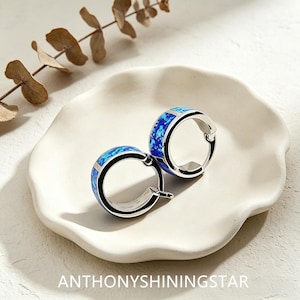 Puede incluir: Un par de pendientes de aro de plata con una incrustación de ópalo azul triturado. Los pendientes tienen un acabado metálico pulido y un mecanismo de cierre de bisagra. El texto ANTHONYSHININGSTAR está impreso en la parte inferior.