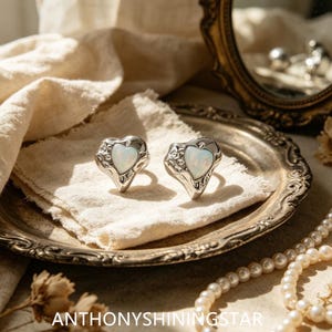 Puede incluir: Dos anillos de tono plateado con una piedra iridiscente en forma de corazón engastada en un marco de metal texturizado de forma orgánica. Los anillos se muestran sobre una tela, con el texto ANTHONYSHININGSTAR visible en la parte inferior.