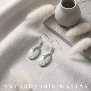 May include: A pair of silver-toned drop earrings featuring oval-shaped pendants with white iridescent inlay and small clear stones. The earrings have curved ear wires and a sunburst-inspired design. The text ANTHONYSHININGSTAR is visible.