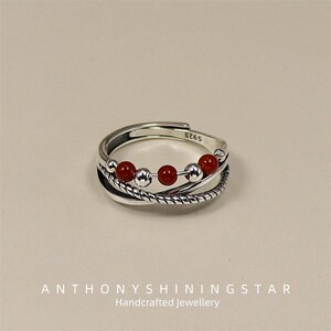 Puede incluir: Un anillo ajustable de plata con un diseño en capas. El anillo presenta cinco cuentas rojas y dos cuentas plateadas. La banda tiene un diseño de cuerda trenzada. El texto "S925" es visible en el anillo. El texto "ANTHONYSHININGSTAR Handcrafted Jewellery" está en la parte inferior.