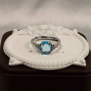 May include: A silver adjustable ring with a large, oval, light blue gemstone. Two smaller, clear gemstones are set either side of the main stone. The ring is displayed on a white decorative tray.