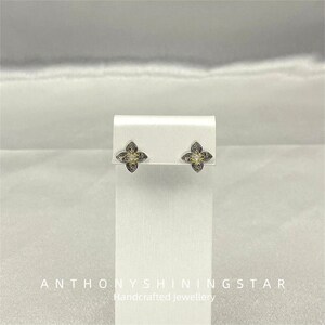 Può includere: Orecchini a lobo argentati con un design a fiore a quattro petali. Ogni orecchino presenta una pietra gialla centrale e dettagli intricati. Sono esposti su un supporto bianco. Il testo "ANTHONYSHININGSTAR Handcrafted Jewellery" è in basso.