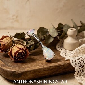 May include: A curved barbell body jewelry piece featuring a small round opal at the top and a larger pear-shaped opal at the bottom, set in a silver-toned metal. The text ANTHONYSHININGSTAR is displayed at the bottom of the image.