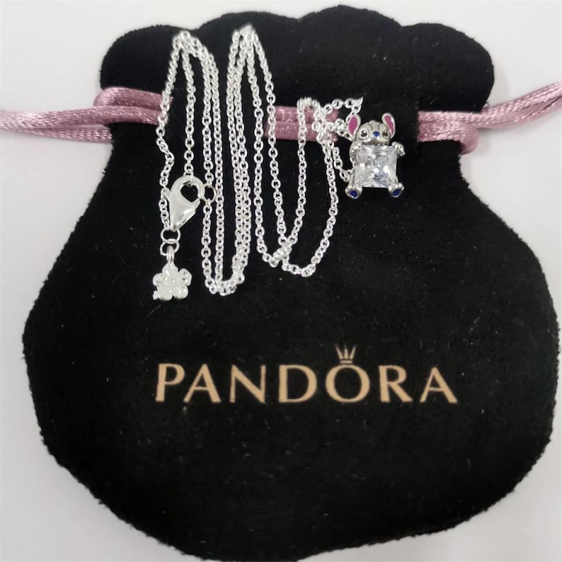 K&ouml;nnte beinhalten: Eine silberne Halskette mit einem Anh&auml;nger einer Zeichentrickfigur, pr&auml;sentiert auf einem schwarzen Samtbeutel mit rosa Kordelzug. Die Halskette hat einen kleinen Blumenanh&auml;nger und einen Karabinerverschluss. Auf dem Beutel ist der Markenname "PANDORA" aufgedruckt.