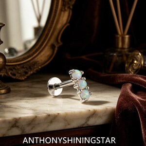 Könnte beinhalten: Ein silberfarbenes Labret-Piercing mit drei runden, schillernden weißen Opalsteinen in einer vertikalen Reihe. Das Schmuckstück hat eine flache, runde Rückplatte und einen geraden Stab. Der Text ANTHONYSHININGSTAR ist unten zu sehen.