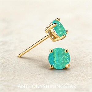 Peut inclure: Une paire de boucles d'oreilles à tige en or avec des pierres rondes bleu sarcelle iridescentes montées sur quatre griffes. Une boucle est vue de côté avec sa tige, l'autre de face. Le texte ANTHONYSHININGSTAR est inscrit en bas de l'image.
