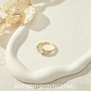May include: A gold-toned open ring featuring a central round white stone flanked by two smaller clear stones on each side. The band has a polished finish and an adjustable opening at the back. The text ANTHONYSHININGSTAR is visible at the bottom.