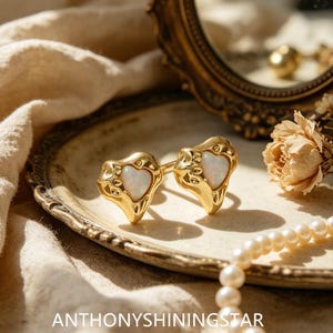 Puede incluir: Un par de pendientes dorados con una piedra de ópalo iridiscente en forma de corazón engastada en un marco de metal texturizado de forma orgánica. El texto ANTHONYSHININGSTAR es visible en la parte inferior, junto a un collar de perlas.