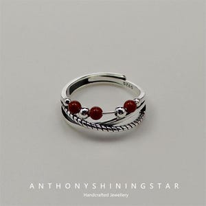 Puede incluir: Un anillo de plata con una banda trenzada y cuentas rojas. El anillo está marcado con el número "S925".
