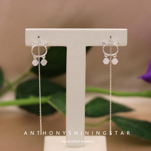 Könnte beinhalten: Silberne Ohrhänger in Katzenform. Jeder Ohrring hat eine Katzenumriss, zwei baumelnde Anhänger und eine lange Kette. Die Ohrringe werden auf einem weißen Ständer vor einem verschwommenen Hintergrund präsentiert. Der Text "ANTHONYSHININGSTAR Handcrafted Jewellery" steht unten.