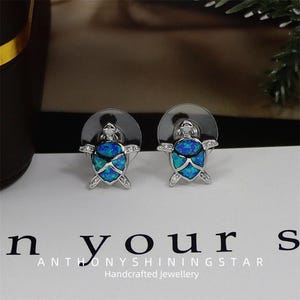 Könnte beinhalten: Silberfarbene Ohrstecker in Form von Schildkröten. Die Ohrringe haben blau-grüne Opal-Schalen und kleine, klare Kristalle. Die Ohrringe liegen auf einer weißen Oberfläche mit dem Text "ANTHONYSHININGSTAR Handcrafted Jewellery."