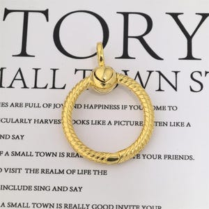 Puede incluir: Un colgante dorado con un diseño en forma de corazón y un anillo circular texturizado. El colgante está suspendido de un pequeño lazo. El fondo presenta la palabra "TORY" y otros textos.