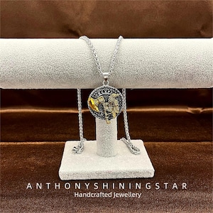May include: A silver-toned necklace with a round pendant featuring a detailed gold and black design. The pendant showcases an eagle and flag motif. The necklace is displayed on a white stand, with the text "ANTHONYSHININGSTAR Handcrafted Jewellery" at the bottom.