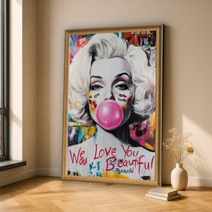 May include: A framed wall art print featuring a stylized portrait of a blonde woman with a bright pink bubblegum bubble. The artwork includes colorful abstract graffiti elements and the handwritten red text We Love You Beautiful.