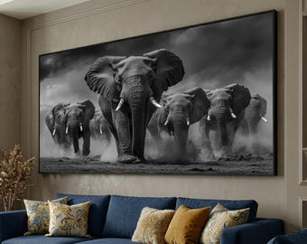 May include: Black and white framed canvas art print featuring a herd of elephants. The elephants are running towards the viewer, kicking up dust. The artwork is displayed above a blue sofa with decorative pillows.