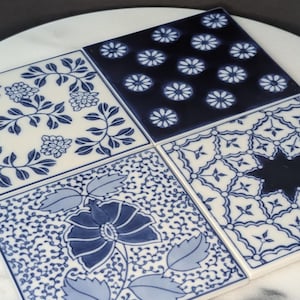 May include: Four square ceramic tiles with blue and white floral and geometric patterns. The tiles are arranged together on a white marble surface. The designs include flowers, leaves, and starbursts.