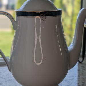 May include: A white porcelain teapot featuring a curved handle, a matching lid with a pointed finial, and a dark teal band near the top rim. The teapot is accented with thin gold trim along the lid, the upper band, and the base of the vessel.