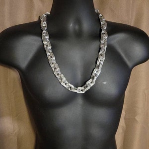 May include: A silver-colored chain necklace with a chunky, interlocking link design. The necklace is displayed on a black mannequin, showcasing its bold and eye-catching style. The chain is made of metal and has a polished finish.