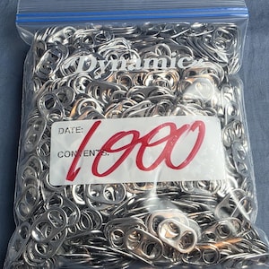May include: A clear plastic resealable bag filled with a large quantity of silver-colored aluminum soda can pull tabs. A white label on the front of the bag features the word Dynamic and the handwritten red text 1000 over the words DATE and CONTENTS.