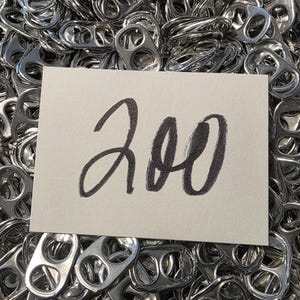 May include: A pile of shiny, silver aluminum can pull tabs. A white card with the number "200" written in black script is placed on top of the tabs. The tabs are densely packed, filling the frame.