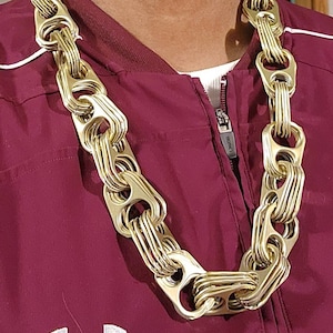 May include: A gold-colored chain necklace made from multiple stacked pull tabs. The necklace is worn by a person wearing a maroon jacket with a white under shirt.