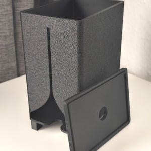 Puede incluir: Un recipiente rectangular negro mate con superficie texturizada, que presenta una ranura vertical en la parte frontal y una tapa rectangular separada con un orificio circular centrado. El recipiente se apoya sobre cuatro patas peque&ntilde;as.