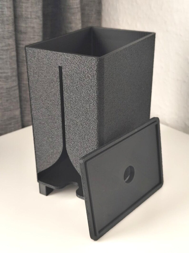Puede incluir: Un recipiente rectangular negro mate con superficie texturizada, que presenta una ranura vertical en la parte frontal y una tapa rectangular separada con un orificio circular centrado. El recipiente se apoya sobre cuatro patas peque&ntilde;as.