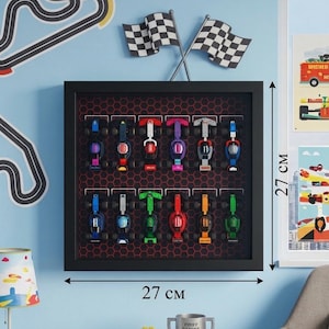 Puede incluir: Una exhibición enmarcada de coches de carreras de juguete coloridos, cada uno con un diseño único. El marco negro mide 27 cm de ancho y 27 cm de alto. También se ven banderas a cuadros y una decoración de pared de circuito.