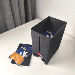 Puede incluir: Un dispensador rectangular negro de textura rugosa con una ranura vertical frontal, que contiene sobres de golosinas para gatos azules y naranjas etiquetados como Jellies. Una tapa negra a juego descansa plana junto al dispensador.