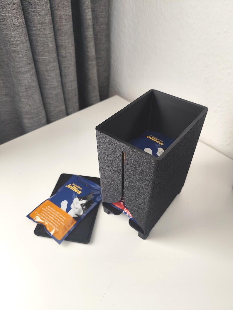 Puede incluir: Un dispensador rectangular negro de textura rugosa con una ranura vertical frontal, que contiene sobres de golosinas para gatos azules y naranjas etiquetados como Jellies. Una tapa negra a juego descansa plana junto al dispensador.