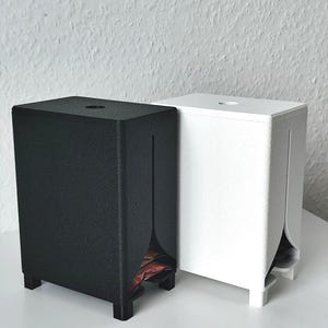 Puede incluir: Dos dispensadores rectangulares, uno negro mate y otro blanco mate, cada uno con una superficie texturizada, cuatro patas peque&ntilde;as, una abertura circular en la parte superior y una ranura vertical en el panel frontal que revela parte del contenido.