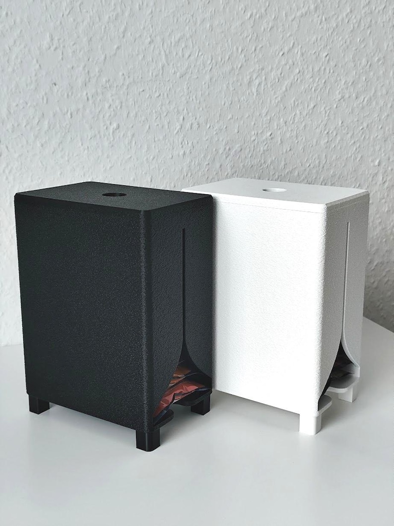 Puede incluir: Dos dispensadores rectangulares, uno negro mate y otro blanco mate, cada uno con una superficie texturizada, cuatro patas peque&ntilde;as, una abertura circular en la parte superior y una ranura vertical en el panel frontal que revela parte del contenido.