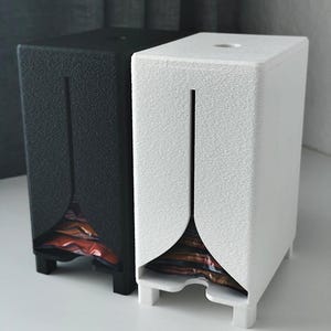 Puede incluir: Dos dispensadores verticales rectangulares, uno negro y otro blanco, con una superficie texturizada, una abertura circular en la parte superior y una ranura vertical frontal que se ensancha en la base para mostrar art&iacute;culos envueltos.