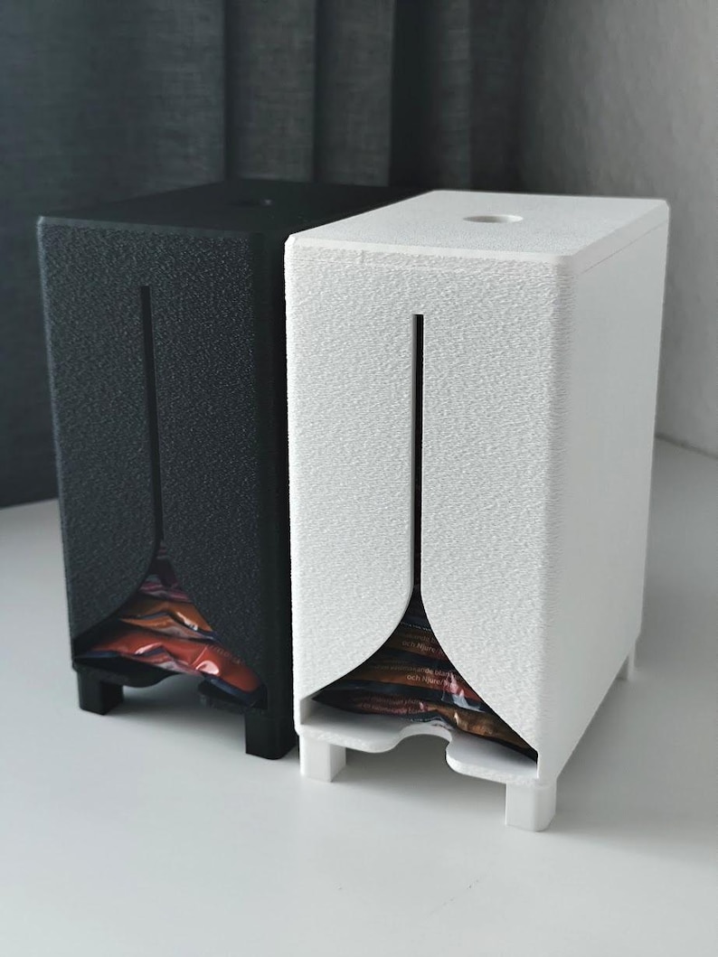 Puede incluir: Dos dispensadores verticales rectangulares, uno negro y otro blanco, con una superficie texturizada, una abertura circular en la parte superior y una ranura vertical frontal que se ensancha en la base para mostrar art&iacute;culos envueltos.