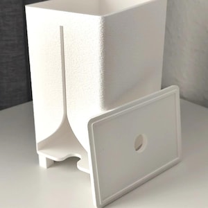 Puede incluir: Un recipiente rectangular blanco con superficie texturizada y una ranura vertical en la parte frontal. Una tapa rectangular blanca separada con un orificio circular centrado se sit&uacute;a frente al recipiente, que se apoya sobre cuatro patas.