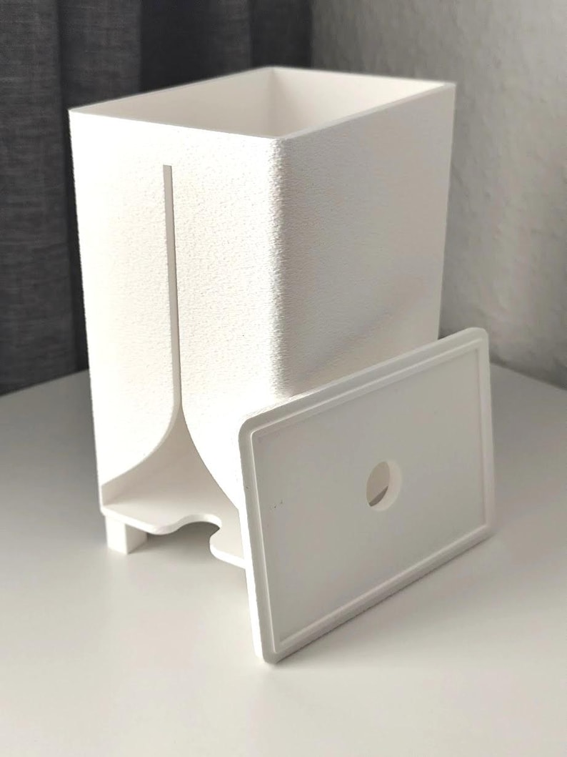 Puede incluir: Un recipiente rectangular blanco con superficie texturizada y una ranura vertical en la parte frontal. Una tapa rectangular blanca separada con un orificio circular centrado se sit&uacute;a frente al recipiente, que se apoya sobre cuatro patas.