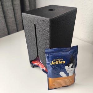 Puede incluir: Un dispensador rectangular de pl&aacute;stico negro texturizado con una ranura vertical en la parte frontal y un orificio circular en la parte superior. Una bolsa azul y naranja de comida para gatos Sensations Jellies con un gato est&aacute; delante.
