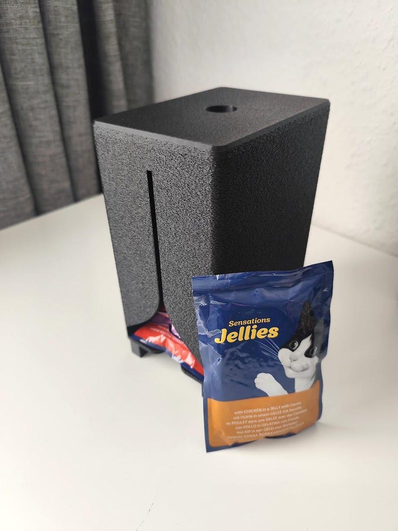 Puede incluir: Un dispensador rectangular de pl&aacute;stico negro texturizado con una ranura vertical en la parte frontal y un orificio circular en la parte superior. Una bolsa azul y naranja de comida para gatos Sensations Jellies con un gato est&aacute; delante.