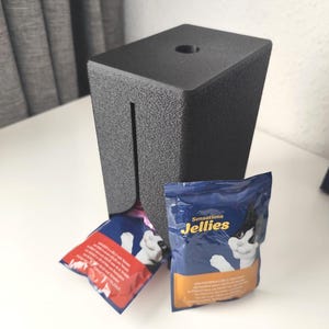 Puede incluir: Un dispensador rectangular de pl&aacute;stico negro texturizado con una ranura vertical y un orificio circular en la parte superior. Se ven dos bolsas de comida para gatos, una roja y otra azul, la azul tiene la etiqueta Sensations Jellies.