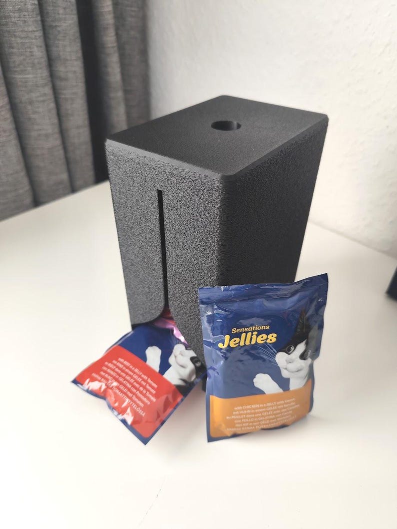 Puede incluir: Un dispensador rectangular de pl&aacute;stico negro texturizado con una ranura vertical y un orificio circular en la parte superior. Se ven dos bolsas de comida para gatos, una roja y otra azul, la azul tiene la etiqueta Sensations Jellies.